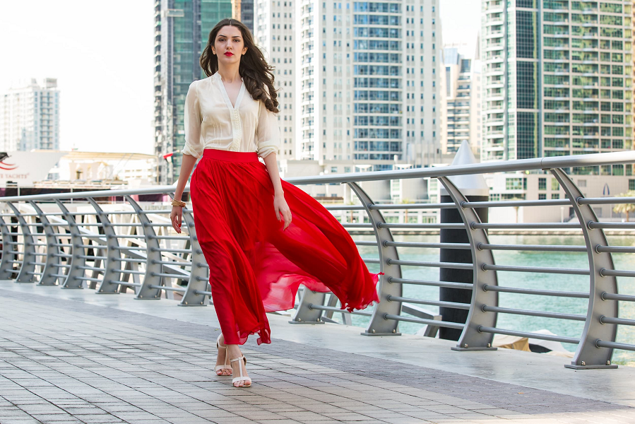 A woman walks along a riverside wearing a long red skirt with strappy sandals.